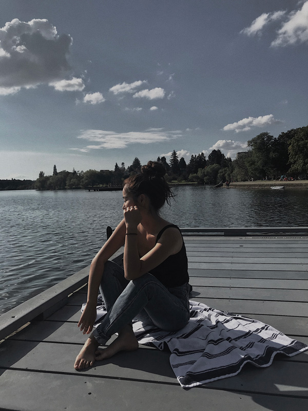 Miwa sitting on a dock by a lake, chin on hand, striped towel beside, trees in the distance.